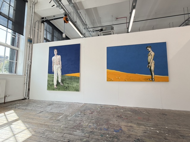 Installation view: two large standing figures on a long wall, lit by windows to the left.